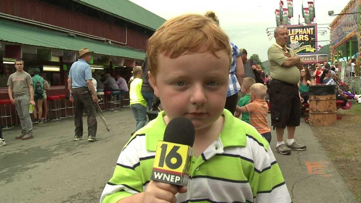 A red-haired child in a lime-green striped shirt holds a WNEP 16 microphone at a busy fairground.
