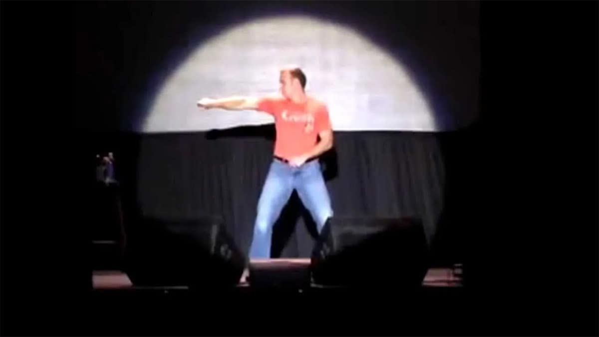 A person in a red shirt and blue jeans performs a dance move on a dark stage, framed by a large circular spotlight and stage equipment.