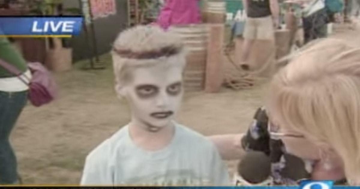 A boy with pale zombie face paint stands beside a reporter holding a microphone during a live outdoor news segment.