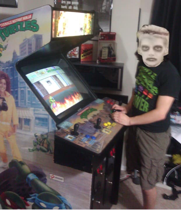A person with a cutout of the meme's pale, dark-eyed zombie face stands beside a Teenage Mutant Ninja Turtles arcade cabinet.