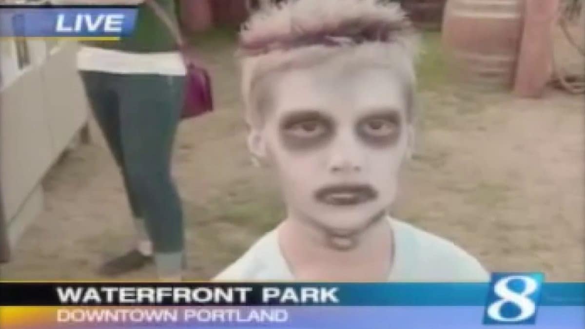 A boy with pale zombie face paint and dark eye makeup looks toward the camera in a local TV frame. The lower third reads “WATERFRONT PARK” and “DOWNTOWN PORTLAND.”