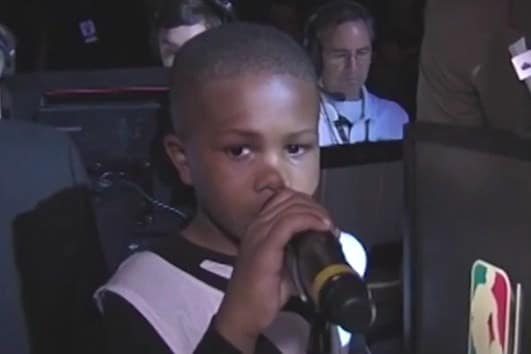 A boy in a black-and-white basketball jersey holds a microphone beside an arena desk during the player-introduction appearance documented in the article.