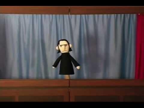 A frame from The Mysterious Ticking Noise shows a stern black-robed felt puppet standing alone on a small wooden stage before blue curtains.