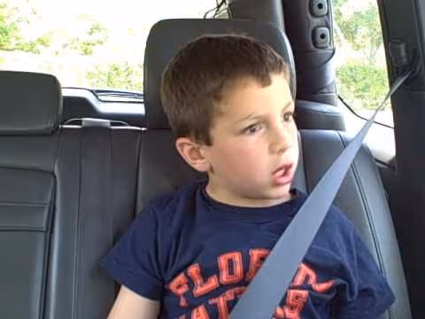 A young boy sits buckled into a car seat and looks to the side, in a frame from the David After Dentist video.