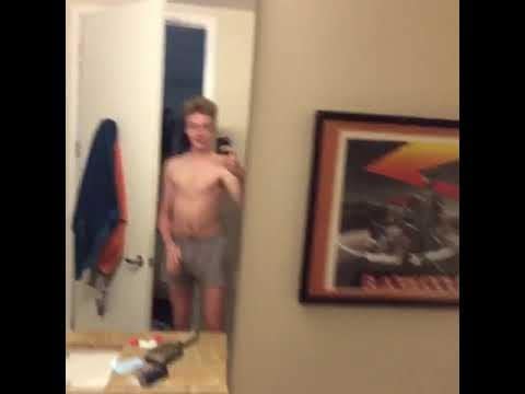 A person in gray shorts films a mirror selfie in a bathroom, with a doorway, counter, and framed wall art visible.